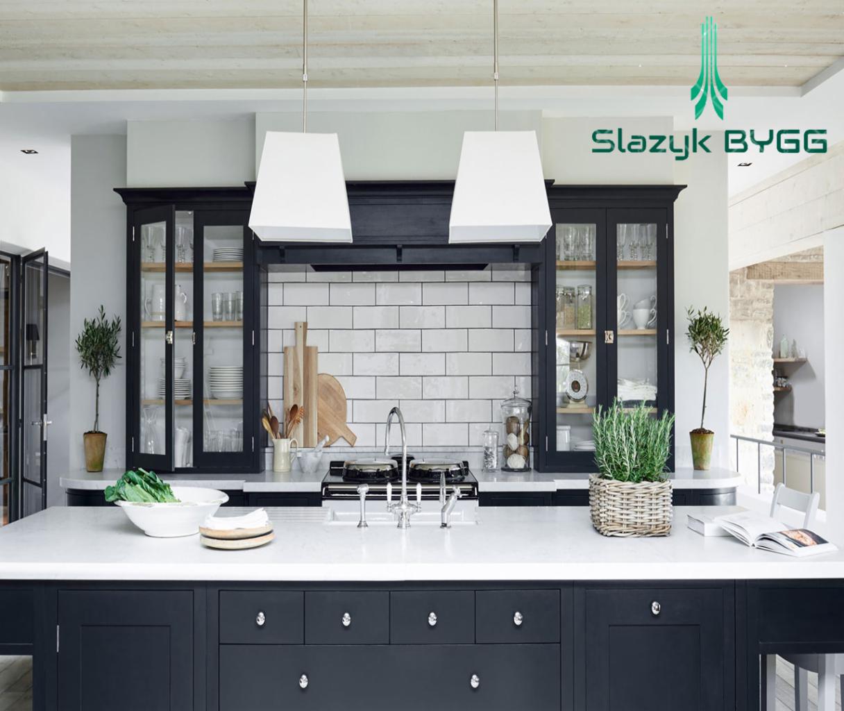Nowoczesna kuchnia z ciemnymi szafkami, białym blatem i płytkami metra na ścianie, oświetlona lampami wiszącymi i z roślinami doniczkowymi na blacie wyspy; logo firmy budowlanej w prawym górnym rogu.