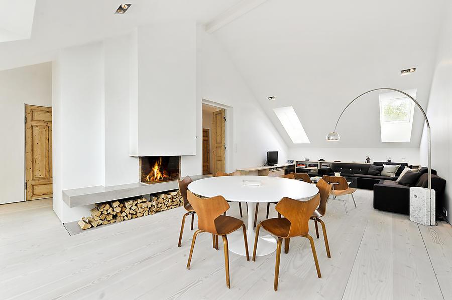 Jasne wnętrze poddasza z białymi ścianami i podłogą, kominkiem z palącym się ogniem i ułożonym drewnem, okrągłym stołem z drewnianymi krzesłami oraz czarną sofą pod oknami dachowymi.