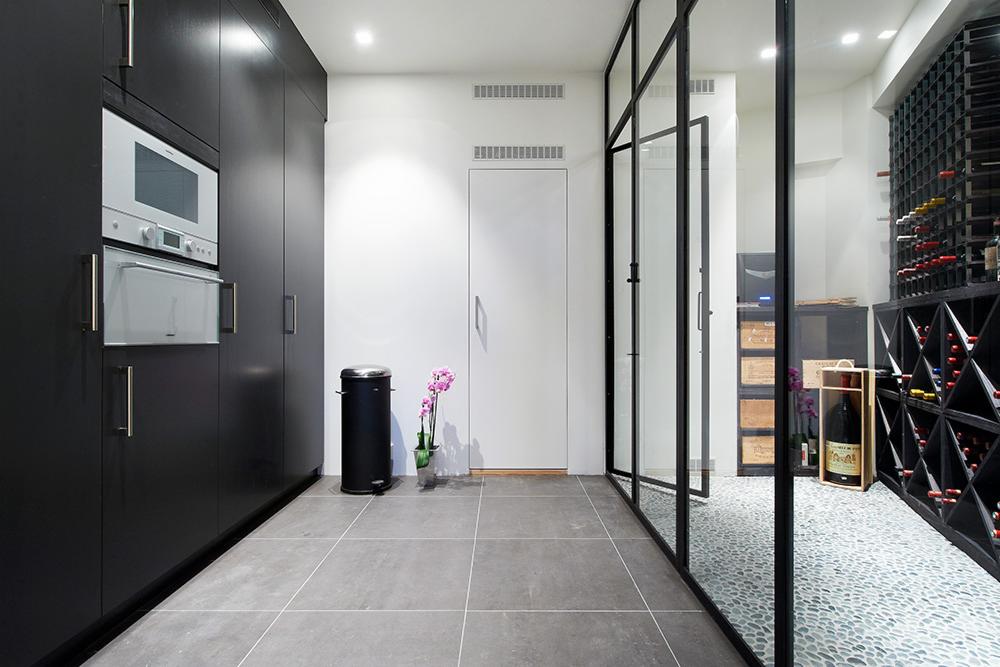 Nowoczesna kuchnia z czarnymi szafkami, wbudowanym piekarnikiem i lodówką, szarą podłogą z płytek, oddzielona szklaną ścianą od piwniczki na wino z kamienną podłogą i regałami na butelki.