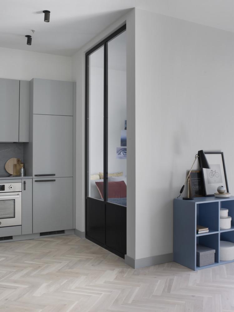 Jasnoszara kuchnia z zabudową, piekarnikiem i szklanymi drzwiami oddzielającymi ją od pokoju z widocznym łóżkiem i szafką nocną z lampą.