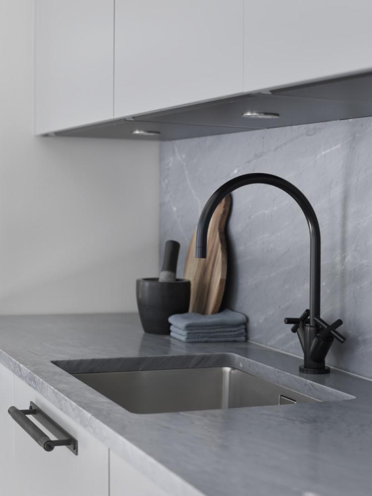 Minimalistyczna kuchnia: stalowy zlew w blacie z szarego marmuru, czarna bateria z krzyżowymi kurkami, moździerz z tłuczkiem, deska do krojenia i szara ściereczka.