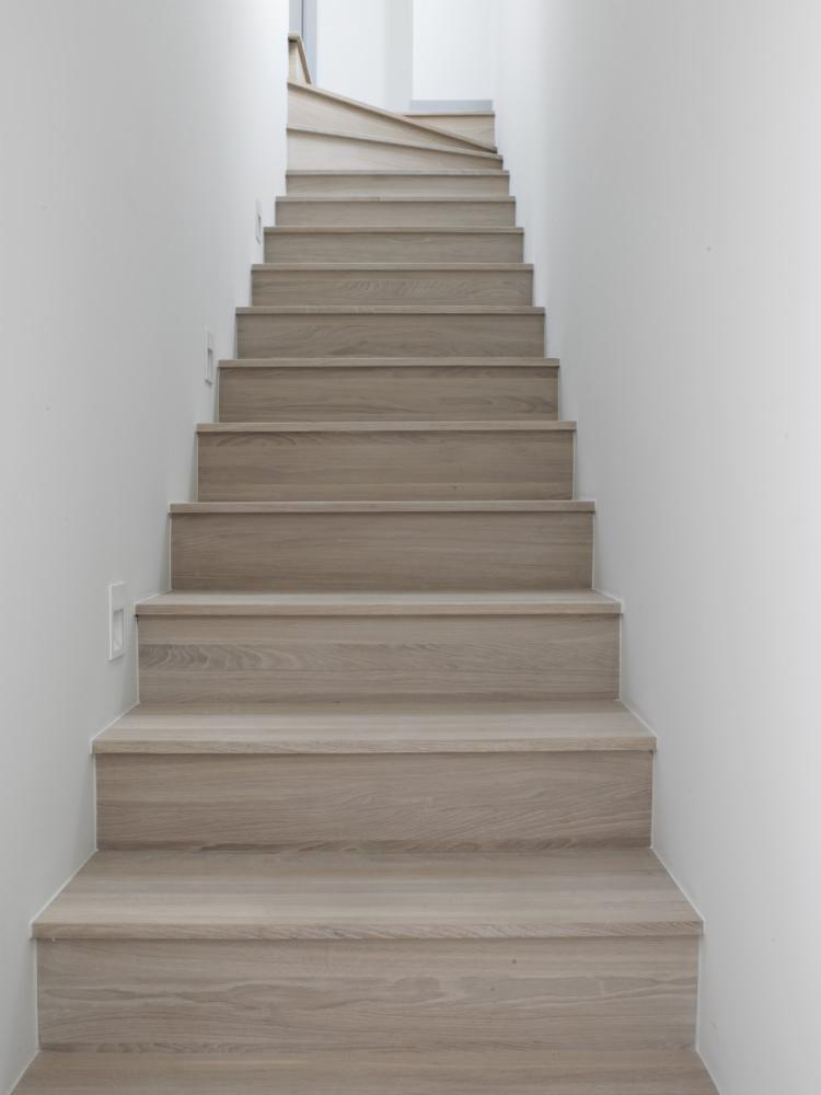 Jasne, drewniane schody prowadzące w górę, otoczone białymi ścianami. Widoczne delikatne oświetlenie punktowe na ścianie.