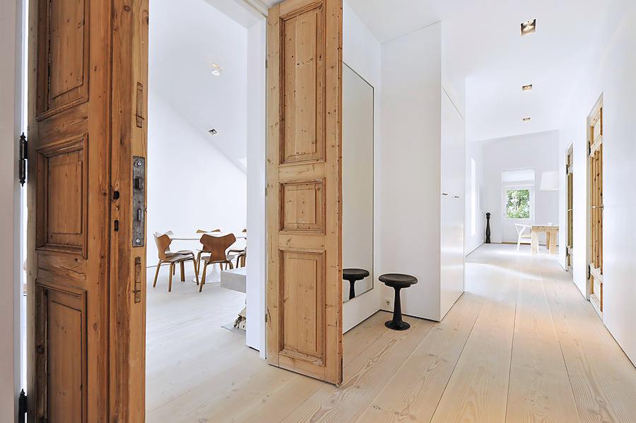 Jasne, przestronne wnętrze z drewnianą podłogą i otwartymi drzwiami z surowego drewna, ukazującymi jadalnię z białym stołem i krzesłami. Nowoczesny design z minimalistycznym wystrojem i naturalnym...