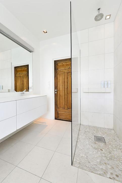 Nowoczesna łazienka z białymi płytkami, szklaną kabiną prysznicową z podłogą wyłożoną otoczakami i drewnianymi drzwiami.