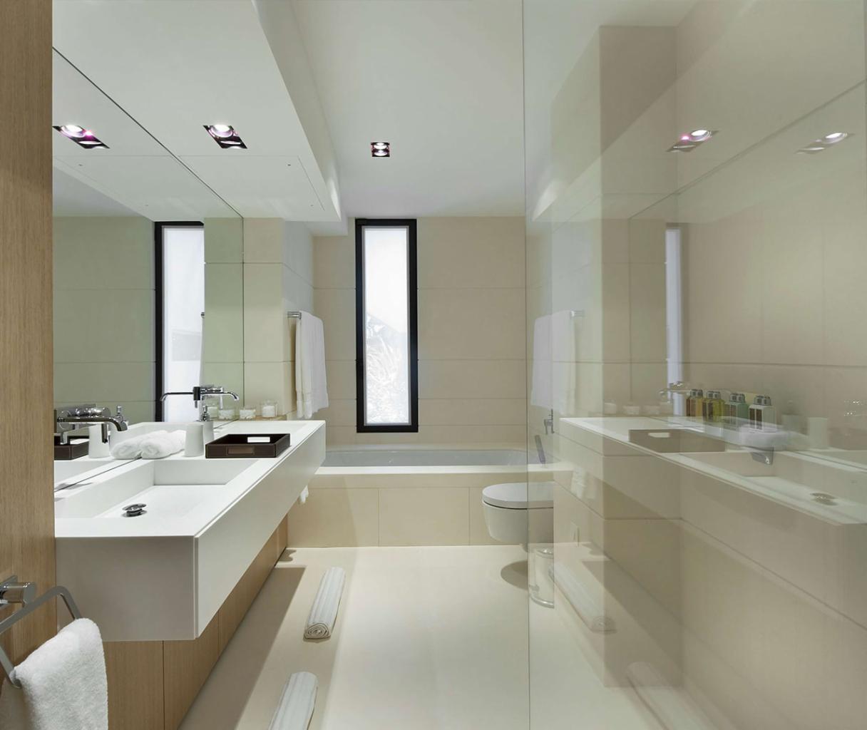 Nowoczesna łazienka w jasnych odcieniach z minimalistyczną umywalką, dużym lustrem, kabiną prysznicową ze szkła i wąskim oknem z widokiem na góry w tle.