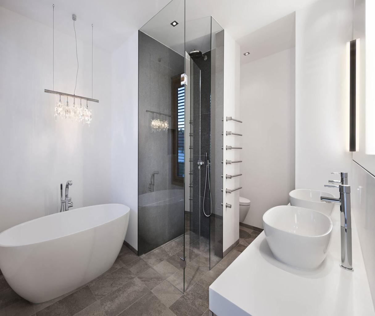Nowoczesna łazienka z wolnostojącą wanną, przeszklonym prysznicem typu walk-in i podwójną umywalką nablatową na białym blacie, oświetlona designerską lampą wiszącą z kryształkami.