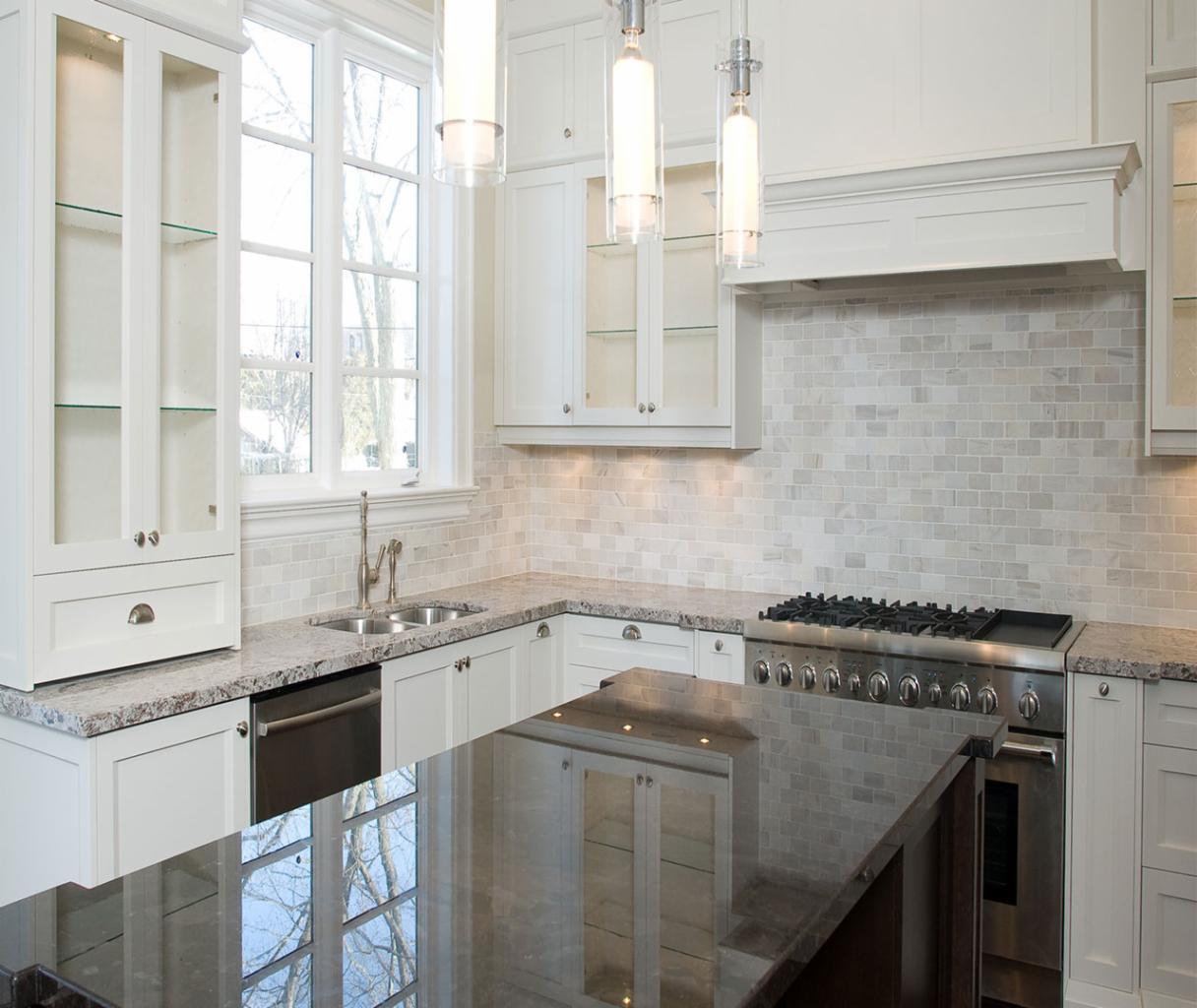 Elegancka, jasna kuchnia z wyspą, białymi szafkami z przeszklonymi witrynami, marmurowym blatem, mozaiką na ścianie, nowoczesnym okapem i lampami wiszącymi.