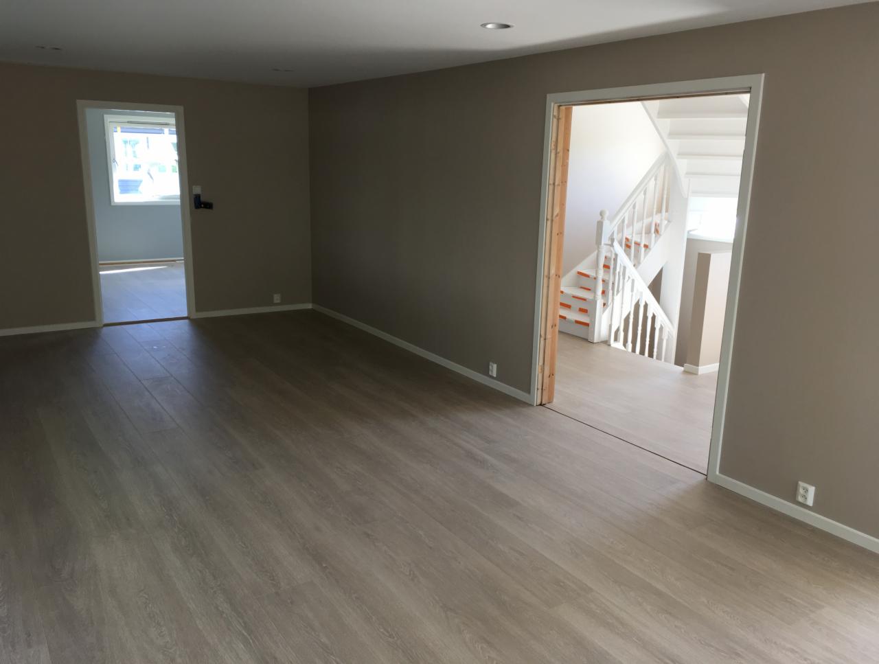 Pusty pokój z panelami podłogowymi w odcieniach szarości, ściany pomalowane na beżowo, widoczne drzwi do innego pomieszczenia i schody z białymi balustradami i pomarańczowymi akcentami na stopniach.