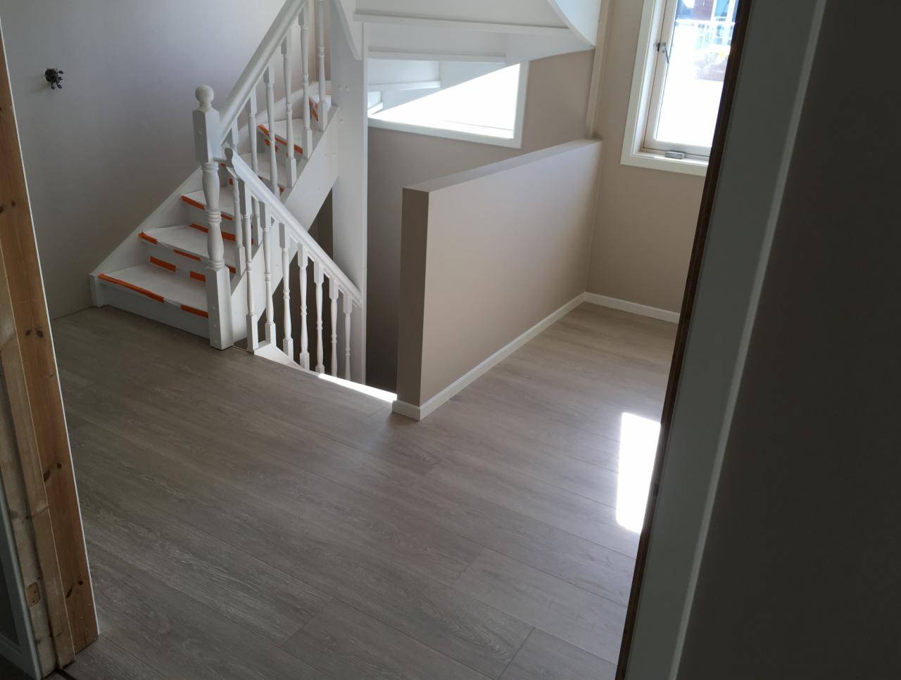 Jasne wnętrze domu w Lommedalen po remoncie: widok na schody z białymi balustradami zabezpieczonymi pomarańczową taśmą malarską, panele podłogowe i świeżo pomalowane ściany w odcieniach beżu.