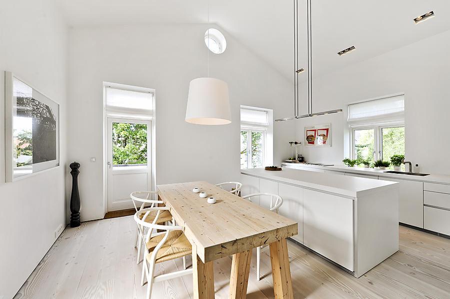 Jasna kuchnia z wysokim sufitem, drewnianym stołem i białymi krzesłami, minimalistyczna zabudowa kuchenna z wyspą, okna z roletami, drewniana podłoga i okrągłe okno w suficie.