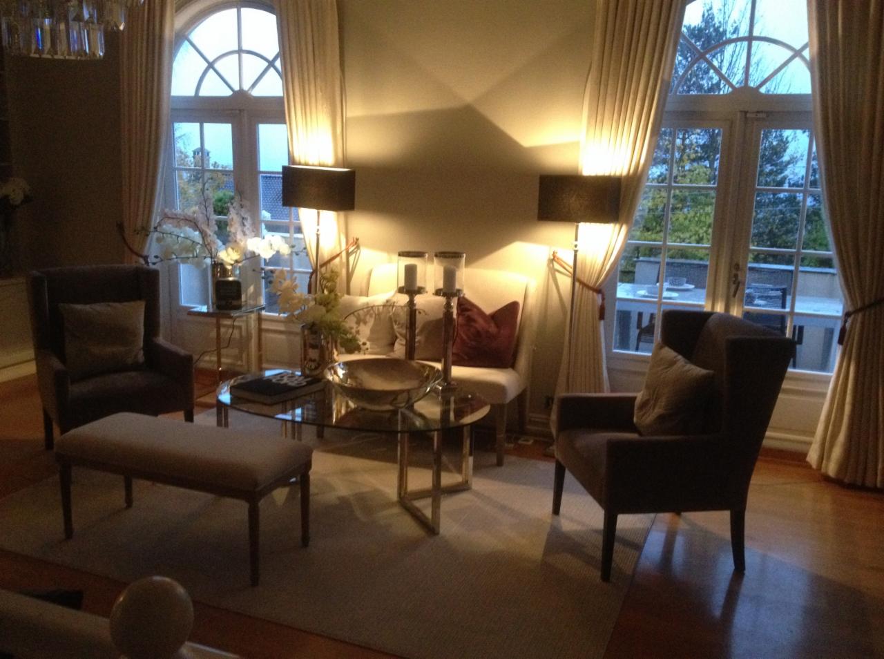 Elegancki salon z dwoma fotelami klubowymi, pufem, szklanym stolikiem kawowym i wysokimi oknami z kremowymi zasłonami oraz lampami podłogowymi z czarnymi abażurami, oświetlony ciepłym światłem.