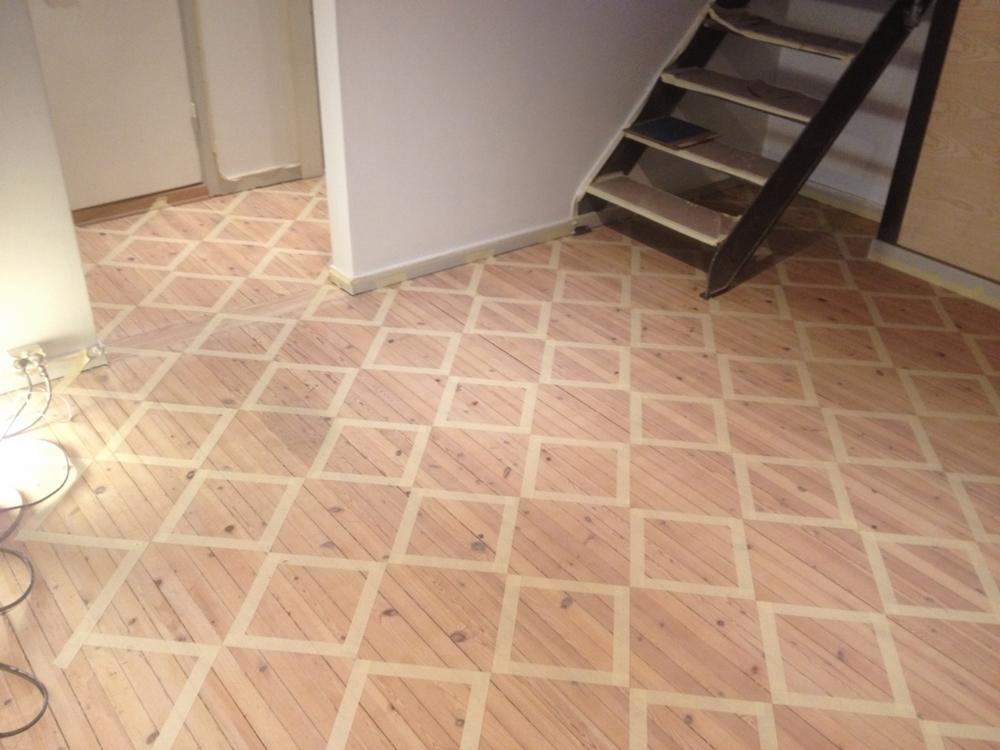 Drewniana podłoga przygotowana do cyklinowania z wyklejonym wzorem w kwadraty przy użyciu taśmy malarskiej, widoczne schody w tle