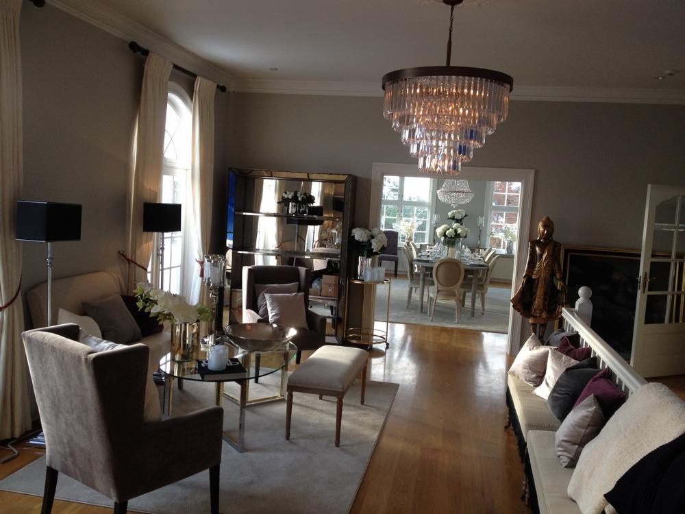 Elegancki salon z parkietem, kryształowym żyrandolem, sofą z poduszkami w odcieniach szarości i fioletu, stolikiem kawowym ze szklanym blatem, lustrzanym regałem i widokiem na jadalnię z dekoracją...