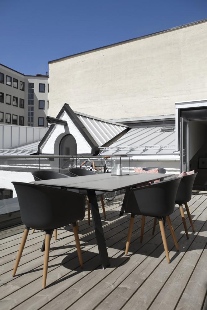 Zadaszony taras z drewnianą podłogą, szarym stołem i czarnymi krzesłami, widok na białe budynki i niebieskie niebo w tle.