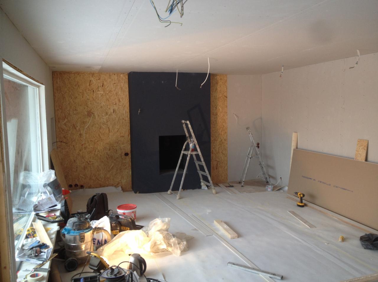 Wnętrze w trakcie prac wykończeniowych: ściany częściowo obłożone płytami OSB, fragment ściany pomalowany na ciemny kolor z otworem, widoczne drabiny, narzędzia i materiały budowlane, folia...