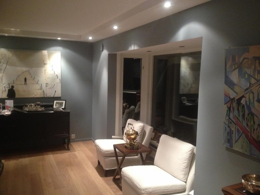 Elegancki salon z drewnianą podłogą, szarymi ścianami, białymi fotelami i nowoczesnym oświetleniem punktowym, ozdobiony obrazami w różnych stylach.