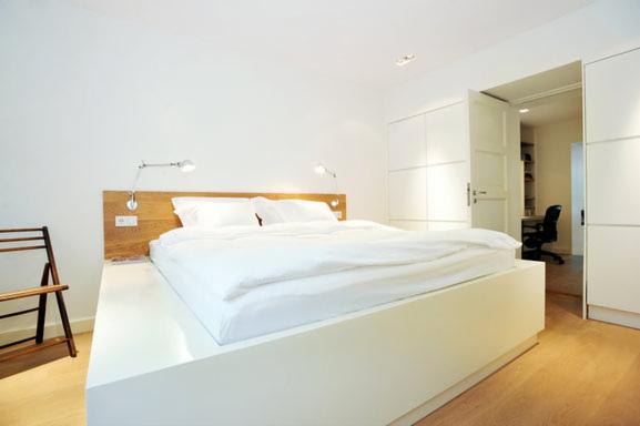 Minimalistyczna sypialnia z białym łóżkiem na podwyższeniu, drewnianym zagłówkiem i lampkami ściennymi, uzupełniona drewnianym krzesłem i szafą w zabudowie.