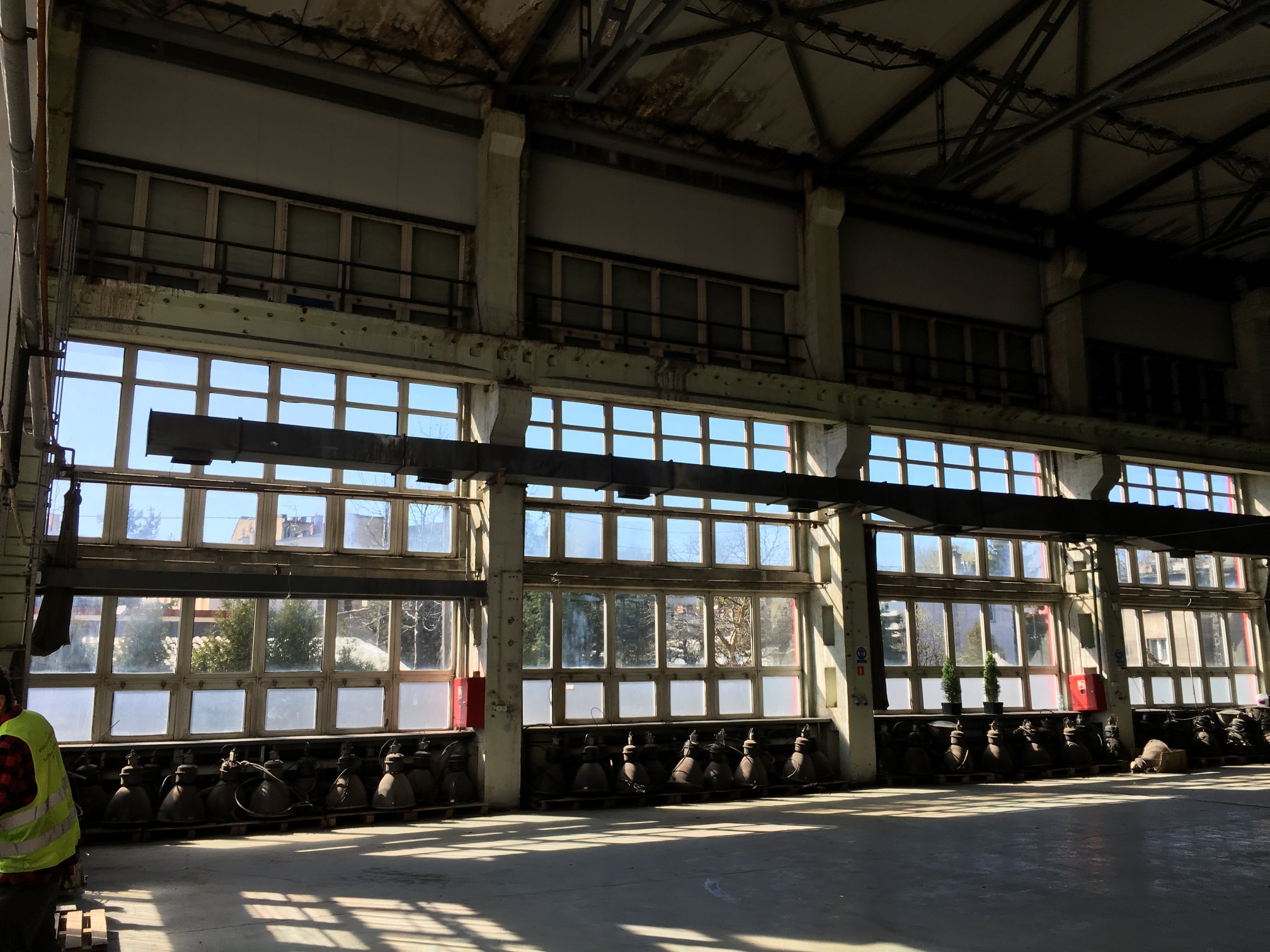 Wnętrze hali przemysłowej z rzędem okien i starymi, wiszącymi lampami na paletach, oświetlonych promieniami słońca wpadającymi przez okna.