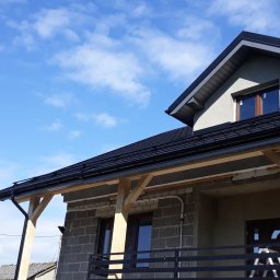 PIO-DACHY - Nowy, czarny dach z rynnami na surowym budynku w trakcie budowy, widoczne drewniane podpory i rusztowania, błękitne niebo w tle.