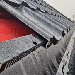 Nowe, czarne orynnowanie z blachy trapezowej, pokryte kroplami deszczu, zamontowane na dachu zabezpieczonym czerwoną membraną dachową, widok z boku, słaba widoczność.