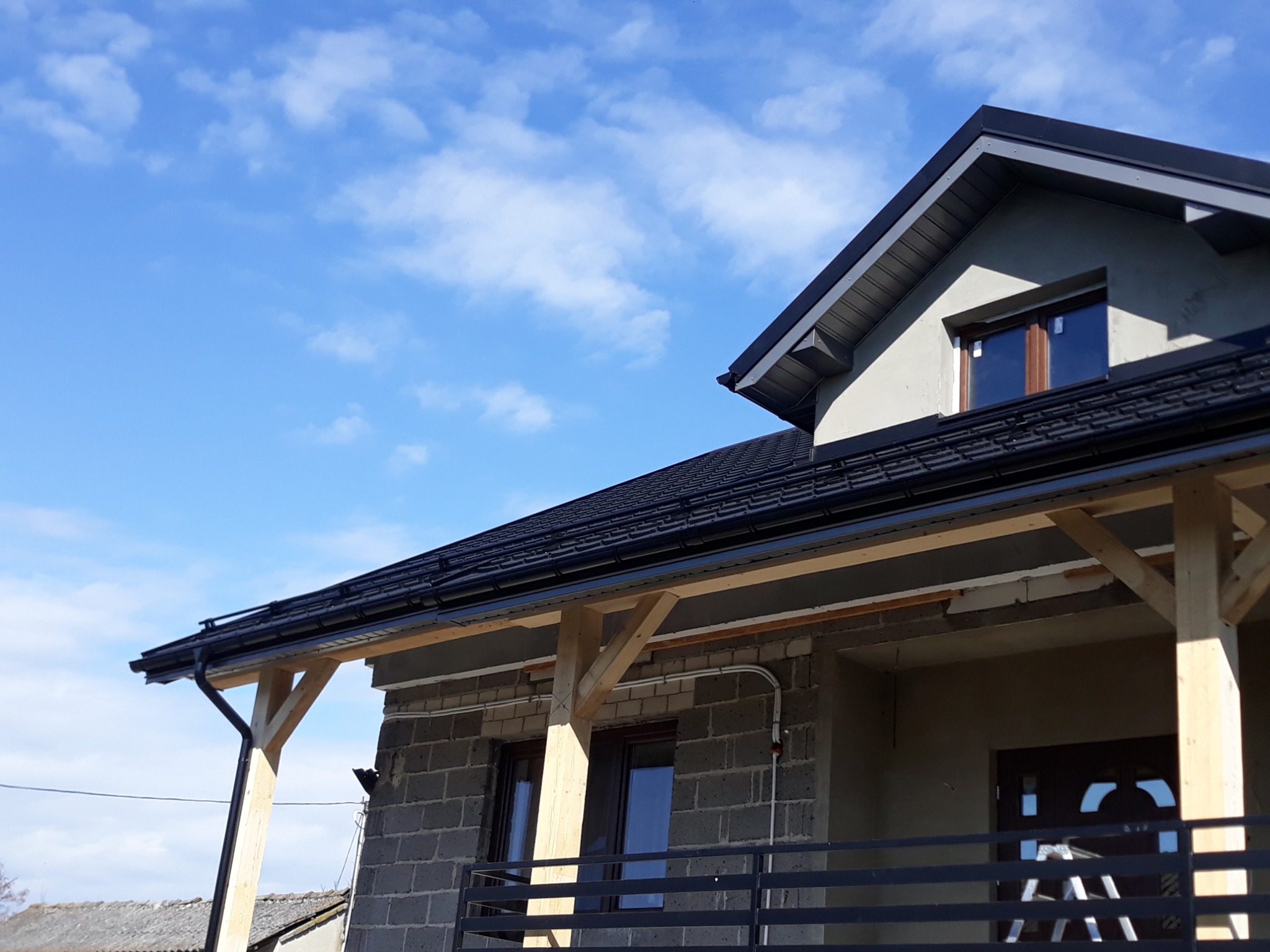 Nowy, czarny dach z rynnami na surowym budynku w trakcie budowy, widoczne drewniane podpory i rusztowania, błękitne niebo w tle.