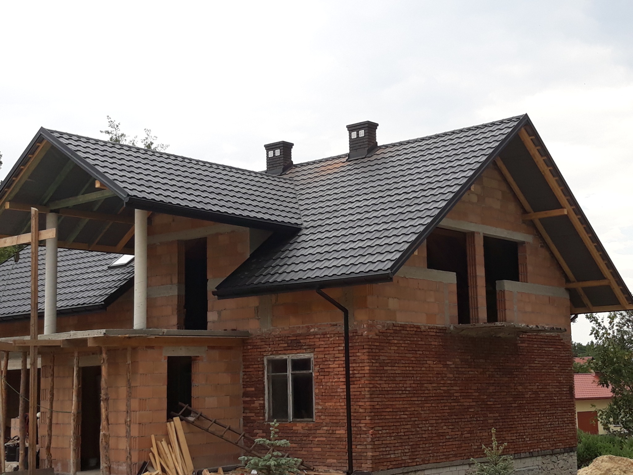 Dom w stanie surowym otwartym z nowym, czarnym dachem z blachodachówki i dwoma kominami, widoczna więźba dachowa i rynny.