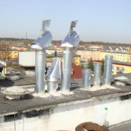 Praca na dachu: mężczyzna w tradycyjnym stroju, w tym cylindrze, montuje metalowe nasady kominowe na tle zabudowy miejskiej