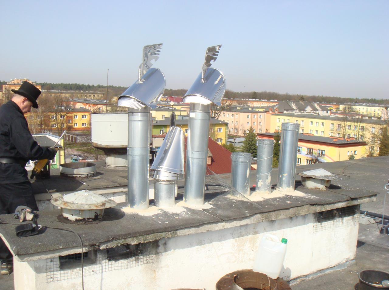 Praca na dachu: mężczyzna w tradycyjnym stroju, w tym cylindrze, montuje metalowe nasady kominowe na tle zabudowy miejskiej