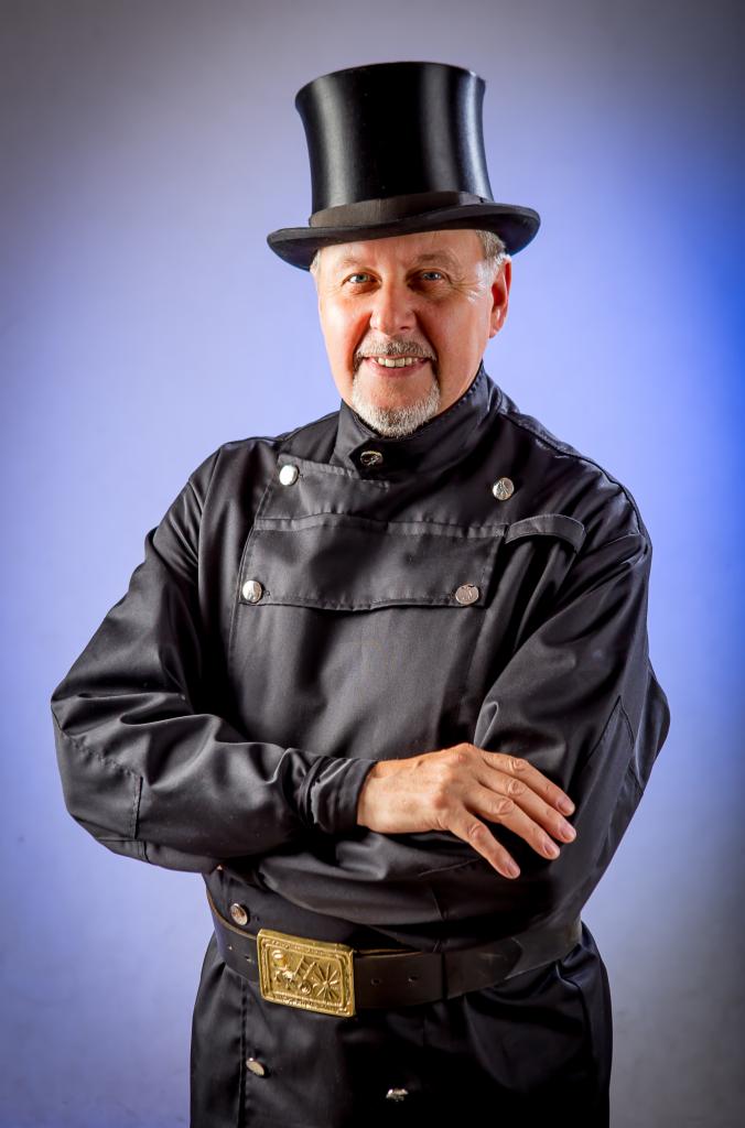 Uśmiechnięty mężczyzna w tradycyjnym stroju z cylindrem, czarnym surducie z srebrnymi guzikami i ozdobną klamrą pasa na tle gradientu niebieskiego.