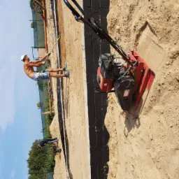 Prace ziemne przy fundamencie: robotnik wyrównuje teren grabiami, obok stoi zagęszczarka wibracyjna, widoczne bloczki fundamentowe pomalowane na czarno.