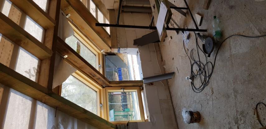 Wnętrze domu w trakcie przebudowy, widoczne okna z drewnianą ramą, ocieplenie ścian, narzędzia budowlane na podłodze i fragment konstrukcji metalowej.