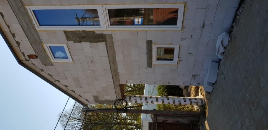 Ściana budynku w trakcie budowy z białych bloczków, widoczne trzy okna z białymi ramami, częściowo otynkowane fragmenty ściany, drewniany słup podtrzymujący konstrukcję i brama w tle.