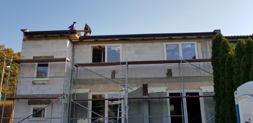 Budowa domu z białego bloczka: dwaj pracownicy na dachu, widoczne rusztowania, wstawione okna, częściowo ocieplona ściana, w tle drzewa.