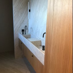 FACH-BUD firma remontowo-malarska - Nowoczesna łazienka z podwójną umywalką z marmuru, czarne baterie i dozownik mydła, drewniana zabudowa szafkowa, ściany wykończone płytami imitującymi marmur, minimalistyczne oświetlenie.