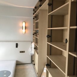 FACH-BUD firma remontowo-malarska - Montaż jasnego, drewnianego regału z półkami i szufladami, widoczne mocowania, oświetlenie ścienne w tle, ochrona podłogi folią.