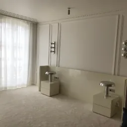 Jasne wnętrze sypialni z dekoracyjnymi panelami ściennymi, białymi zasłonami i kremową wykładziną dywanową. Dwie szafki nocne z lampkami w stylu retro po bokach niskiego zagłówka.