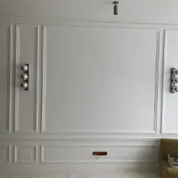 Biała ściana z dekoracyjnymi panelami, oświetleniem ściennym i listwą przypodłogową, widoczne niedokończone gniazdko elektryczne i fragment sofy z tekstyliami.