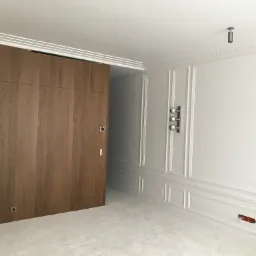 Nowoczesne wnętrze z drewnianą przesuwną ścianą, sztukaterią na ścianach i suficie, oraz minimalistycznym oświetleniem punktowym.