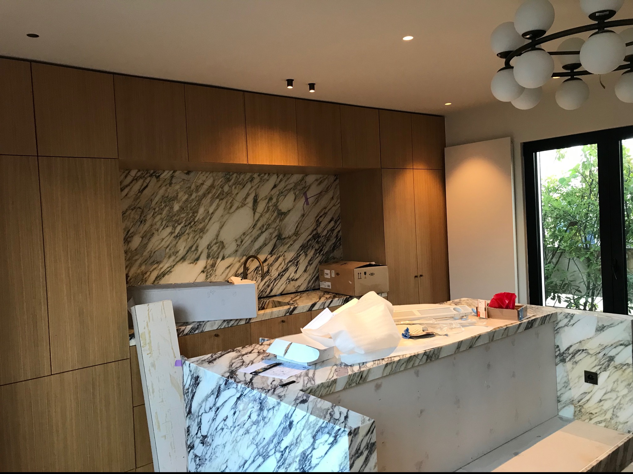 Wnętrze kuchni w trakcie remontu, widoczne marmurowe blaty, drewniane szafki, kartony i pozostałości po pracach wykończeniowych, okno z widokiem na zieleń.