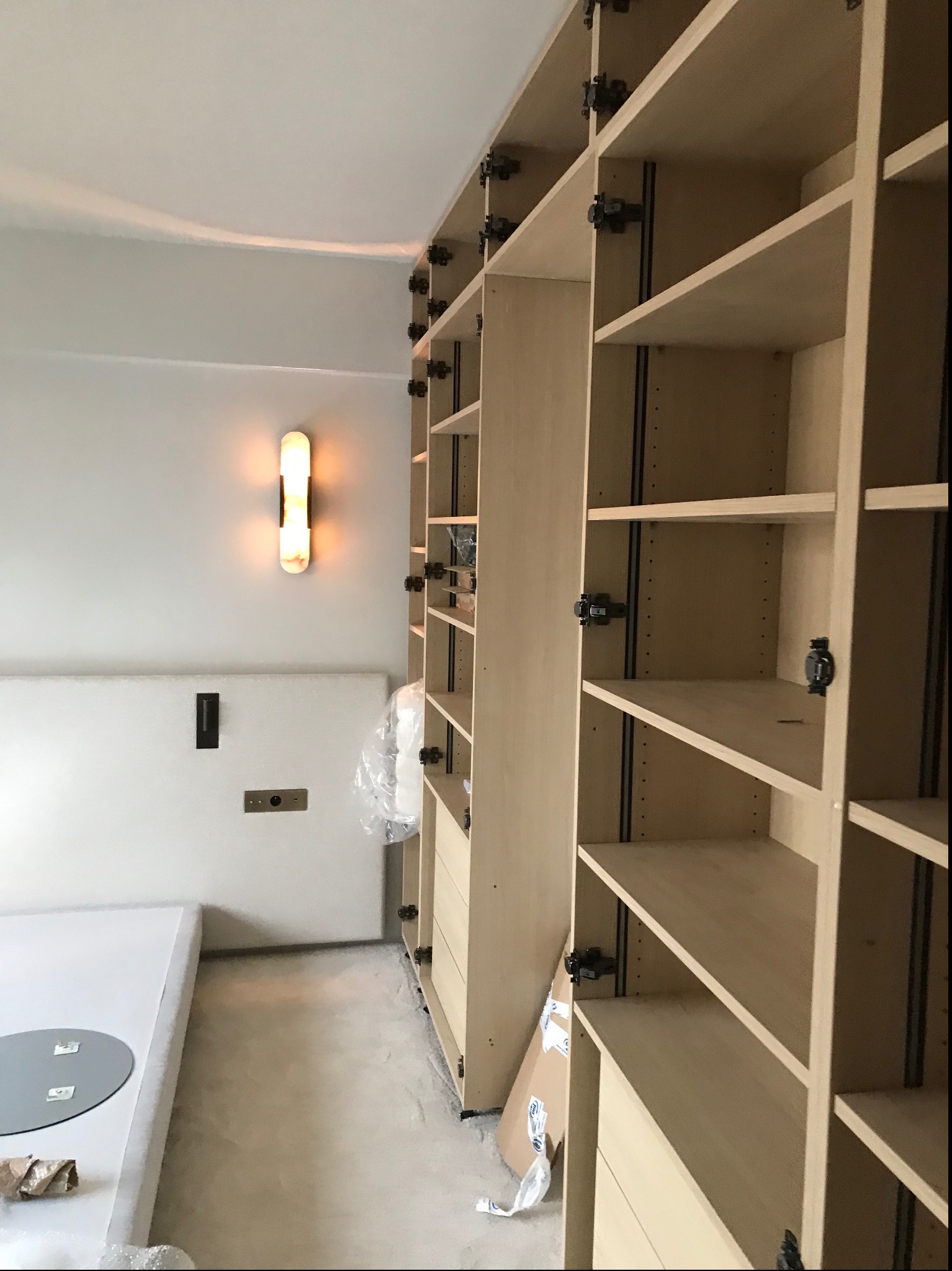 Montaż jasnego, drewnianego regału z półkami i szufladami, widoczne mocowania, oświetlenie ścienne w tle, ochrona podłogi folią.