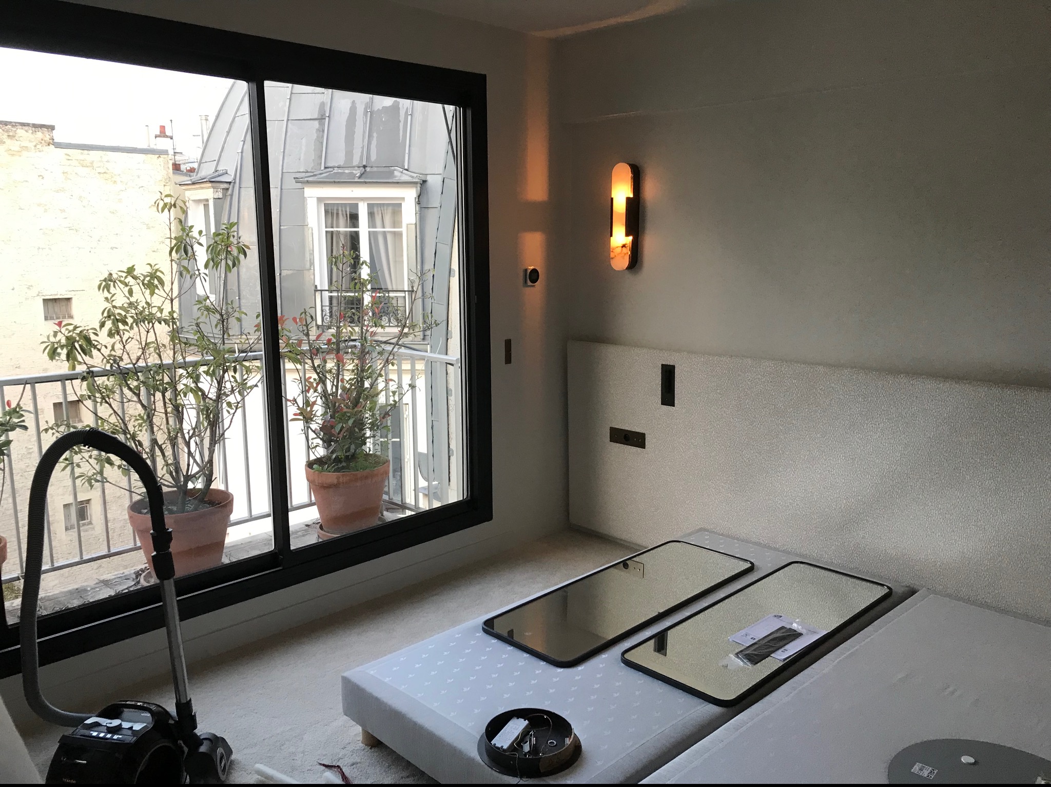 Nowoczesne wnętrze w trakcie remontu: widoczny odkurzacz Miele, przygotowane lustra do montażu na tle okna z widokiem na budynki z blachy falistej i rośliny w donicach.