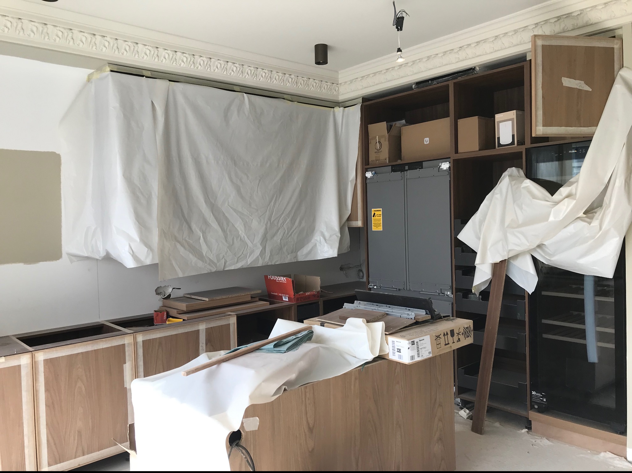 Wnętrze kuchni w trakcie montażu: częściowo zamontowane szafki z okleiną drewnopodobną, zabezpieczone folią malarską ściany i sufit z ozdobną sztukaterią, widoczne kartony i narzędzia.