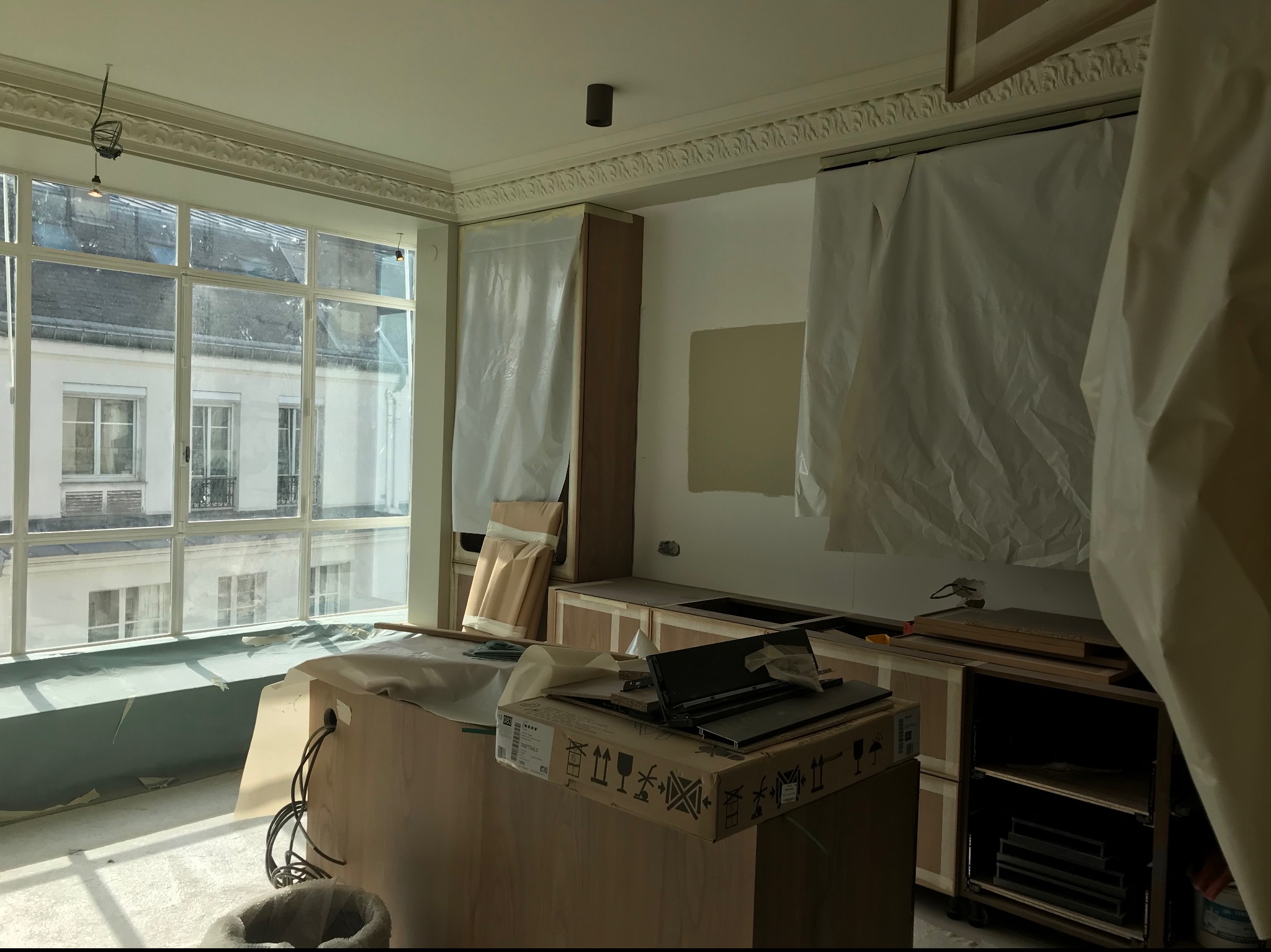 Remont kuchni w kamienicy: okno z widokiem na budynki, zabezpieczone folią meble kuchenne w trakcie montażu, karton z elementami, ślady po malowaniu na ścianie.