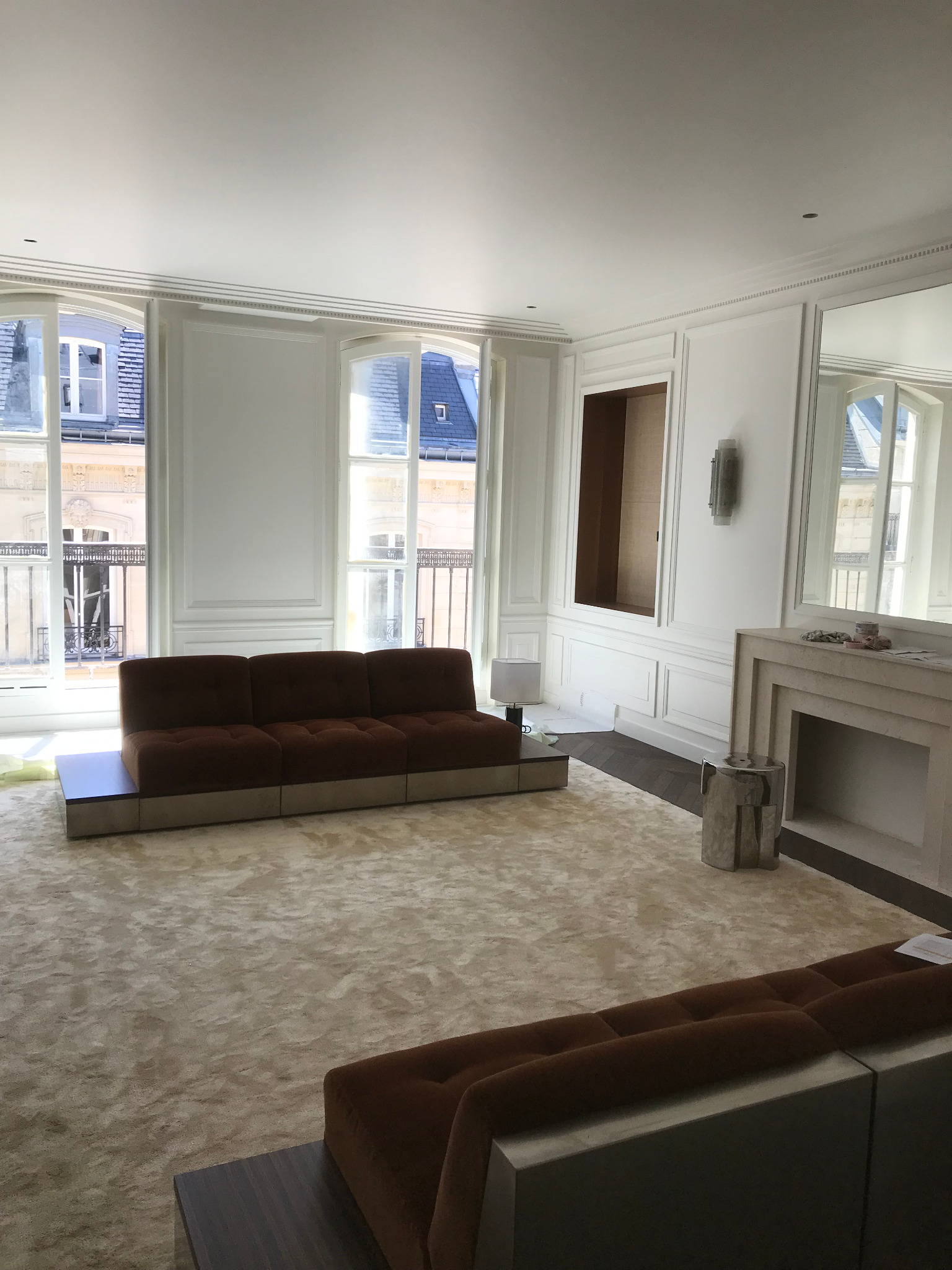 Jasne wnętrze po remoncie z kremowym dywanem, brązową sofą i białymi ścianami z dekoracyjnymi panelami, widoczne okna z widokiem na miasto.