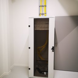 Usługi Elektryczno Budowlane Leszek Drewczyński - Biała szafa rack z żółtymi kablami wewnątrz, widoczne kable w korytach na suficie, szare drzwi obok. Minimalistyczne wnętrze, jasne oświetlenie.