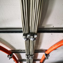 Usługi Elektryczno Budowlane Leszek Drewczyński - Zamocowane kable elektryczne w peszlach i wiązkach, prowadzone pod sufitem. Widoczne metalowe uchwyty mocujące i fragmenty instalacji.