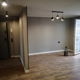 Usługi Elektryczno Budowlane Leszek Drewczyński - Szare wnętrze po remoncie: drewnopodobna podłoga, ściany w odcieniach szarości, nowoczesne oświetlenie sufitowe i otwarte drzwi balkonowe wpuszczające światło.