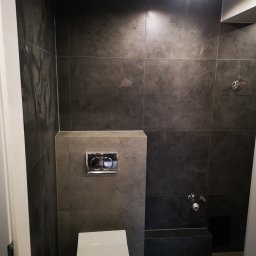 Usługi Elektryczno Budowlane Leszek Drewczyński - Nowoczesna łazienka z ciemnymi, grafitowymi płytkami, wiszącą toaletą i chromowanym przyciskiem spłukiwania. Widoczne przyłącza wodne i fragment instalacji elektrycznej.