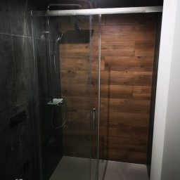 Usługi Elektryczno Budowlane Leszek Drewczyński - Nowoczesna kabina prysznicowa z drewnopodobnymi płytkami na ścianie, szklane drzwi, czarne płytki po lewej stronie, minimalistyczny design.