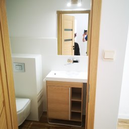 Usługi Elektryczno Budowlane Leszek Drewczyński - Nowoczesna łazienka po remoncie: białe płytki z subtelnym wzorem, drewniana szafka pod umywalką z otwartymi półkami, podłoga imitująca drewno, widok zza otwartych drzwi.